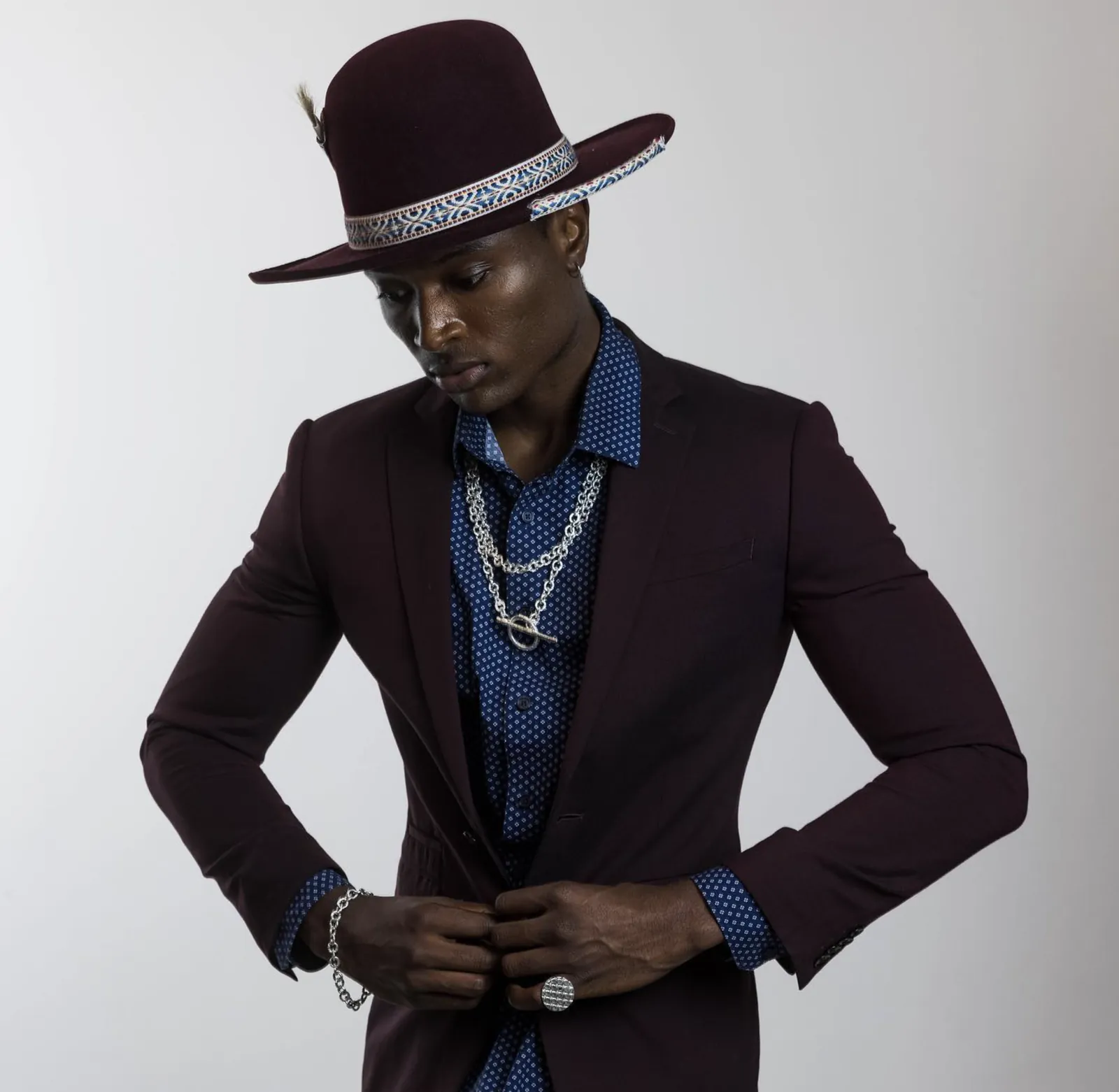 Model in a burgundy suit and hat wearing layered chains with a toggle clasp, a chain bracelet and a textured ring